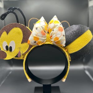 May include: Yellow and black bee-themed Minnie Mouse ears with a honeycomb bow. The ears are made of felt and have a 3D bee design. The bow is made of fabric and has a honeycomb pattern.