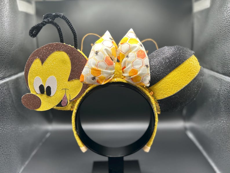 May include: Yellow and black bee-themed Minnie Mouse ears with a honeycomb bow. The ears are made of felt and have a 3D bee design. The bow is made of fabric and has a honeycomb pattern.