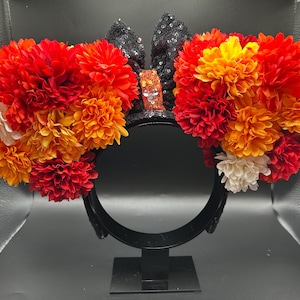 May include: A black headband featuring two circular ears covered in black sequins. Each ear is adorned with a dense arrangement of artificial flowers in shades of red, orange, and white. A small orange sequined bow sits between the ears.