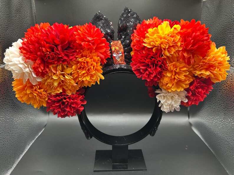 May include: A black headband featuring two circular ears covered in black sequins. Each ear is adorned with a dense arrangement of artificial flowers in shades of red, orange, and white. A small orange sequined bow sits between the ears.