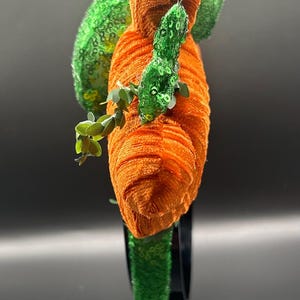 May include: An orange and green headpiece with a unique design. The headpiece features an orange, textured, cylindrical shape, with green sequined accents and leaf-like details. Small green leaves are attached to the orange cylinder.
