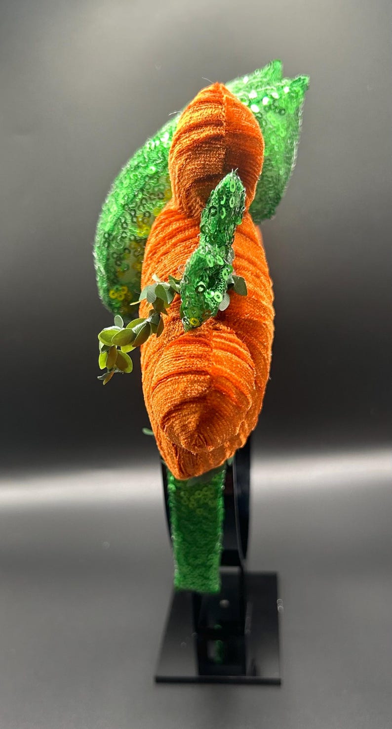 May include: An orange and green headpiece with a unique design. The headpiece features an orange, textured, cylindrical shape, with green sequined accents and leaf-like details. Small green leaves are attached to the orange cylinder.