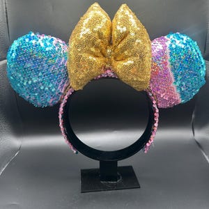 May include: A headband featuring two circular ears covered in iridescent blue, purple, and pink sequins. A large, central bow made of shimmering gold sequins sits between the ears. The headband frame is black and lined with small pink sequins.