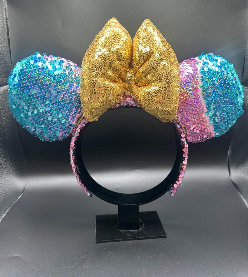 May include: A headband featuring two circular ears covered in iridescent blue, purple, and pink sequins. A large, central bow made of shimmering gold sequins sits between the ears. The headband frame is black and lined with small pink sequins.