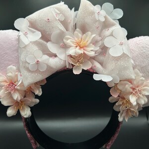 Op de afbeelding: Een haarband met twee lichtroze pluche oren en een grote, glinsterende, lichtroze stoffen strik in het midden. De strik en de basis van de oren zijn versierd met meerdere witte en lichtroze kunstbloemen en kleine kraaltjes.
