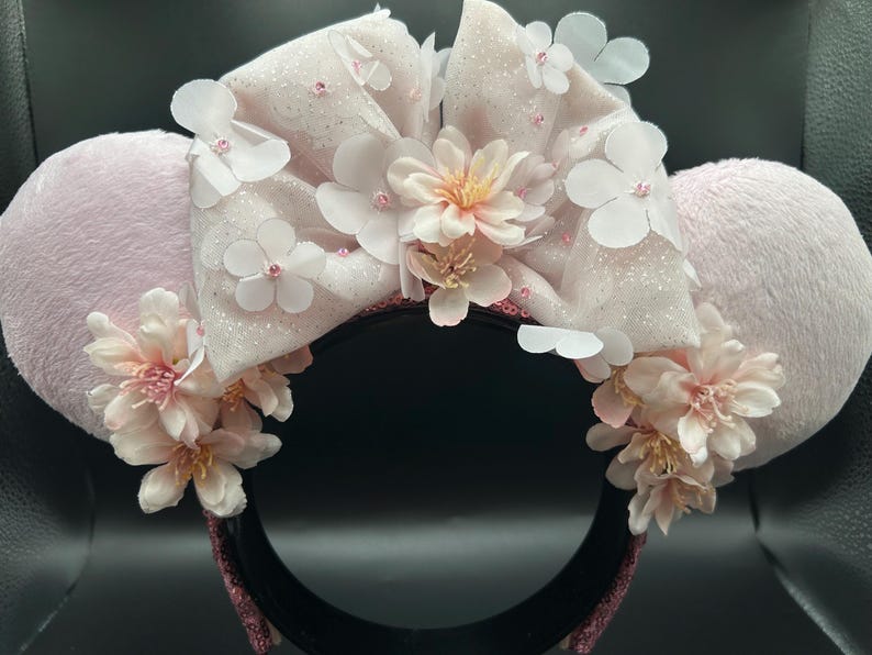 Op de afbeelding: Een haarband met twee lichtroze pluche oren en een grote, glinsterende, lichtroze stoffen strik in het midden. De strik en de basis van de oren zijn versierd met meerdere witte en lichtroze kunstbloemen en kleine kraaltjes.