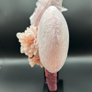May include: A light pink headband with a plush, oval-shaped centre. The headband is adorned with clusters of peach-coloured flowers and delicate white floral accents. The base of the headband is a deep pink colour, and it is displayed on a black stand.