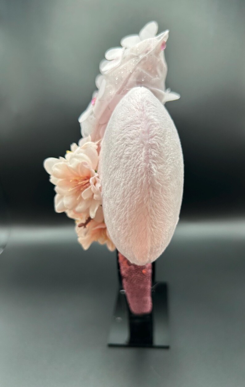 May include: A light pink headband with a plush, oval-shaped centre. The headband is adorned with clusters of peach-coloured flowers and delicate white floral accents. The base of the headband is a deep pink colour, and it is displayed on a black stand.