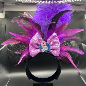 May include: A headband featuring black sequined ears, a central light purple sequined bow, and a vibrant arrangement of purple and magenta feathers. A circular enamel pin depicting a character with long hair is centered on the bow.