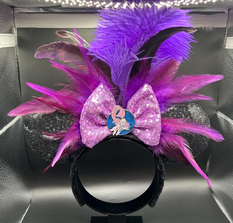 May include: A headband featuring black sequined ears, a central light purple sequined bow, and a vibrant arrangement of purple and magenta feathers. A circular enamel pin depicting a character with long hair is centered on the bow.