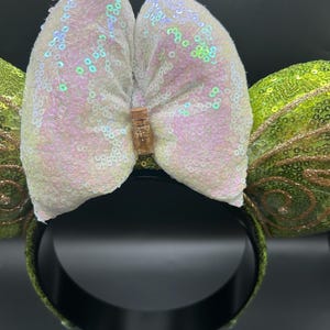 Op de afbeelding: Een haarband met twee ronde oren bedekt met limoengroene lovertjes en gouden glitterwervelpatronen. Tussen de oren zit een grote, iriserende witte strik van lovertjes, in het midden voorzien van een miniatuurflesje gevuld met gouden glitters.