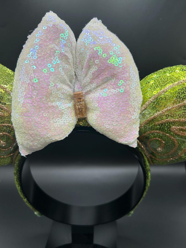 Op de afbeelding: Een haarband met twee ronde oren bedekt met limoengroene lovertjes en gouden glitterwervelpatronen. Tussen de oren zit een grote, iriserende witte strik van lovertjes, in het midden voorzien van een miniatuurflesje gevuld met gouden glitters.
