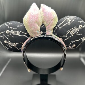 May include: Black headband with two round ears patterned with white skeletons on a black background. The center features two sequined ears in a light pink color. The headband is decorated with sequins. A fun accessory for Halloween or themed events.
