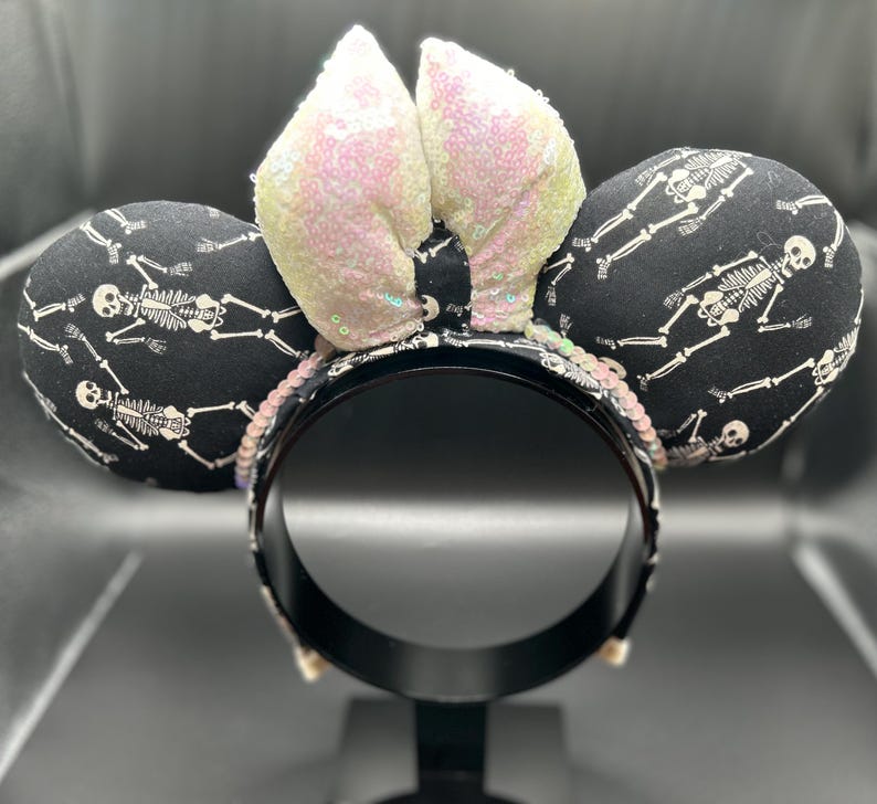 May include: Black headband with two round ears patterned with white skeletons on a black background. The center features two sequined ears in a light pink color. The headband is decorated with sequins. A fun accessory for Halloween or themed events.