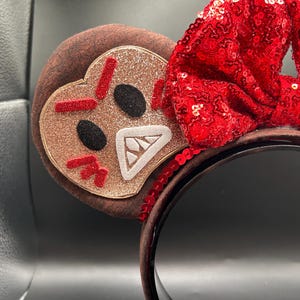 May include: A headband featuring two round ears covered in brown fabric. One ear displays a glittery tan gingerbread character with an angry expression, featuring red stitched eyebrows, black eyes, and a white mouth with teeth. A large red sequined bow sits between the ears.