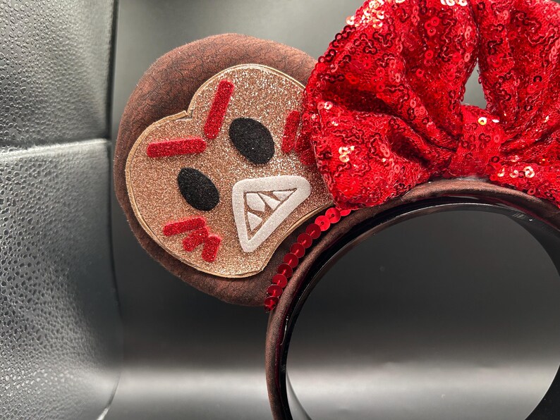 May include: A headband featuring two round ears covered in brown fabric. One ear displays a glittery tan gingerbread character with an angry expression, featuring red stitched eyebrows, black eyes, and a white mouth with teeth. A large red sequined bow sits between the ears.