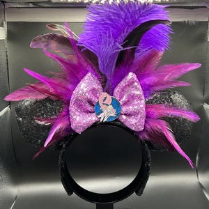 May include: A headband featuring two black sequined ears, a central bright purple sequined bow, and a cluster of vibrant purple and magenta feathers. A circular enamel pin depicting a cartoon character with long hair is centered on the bow.