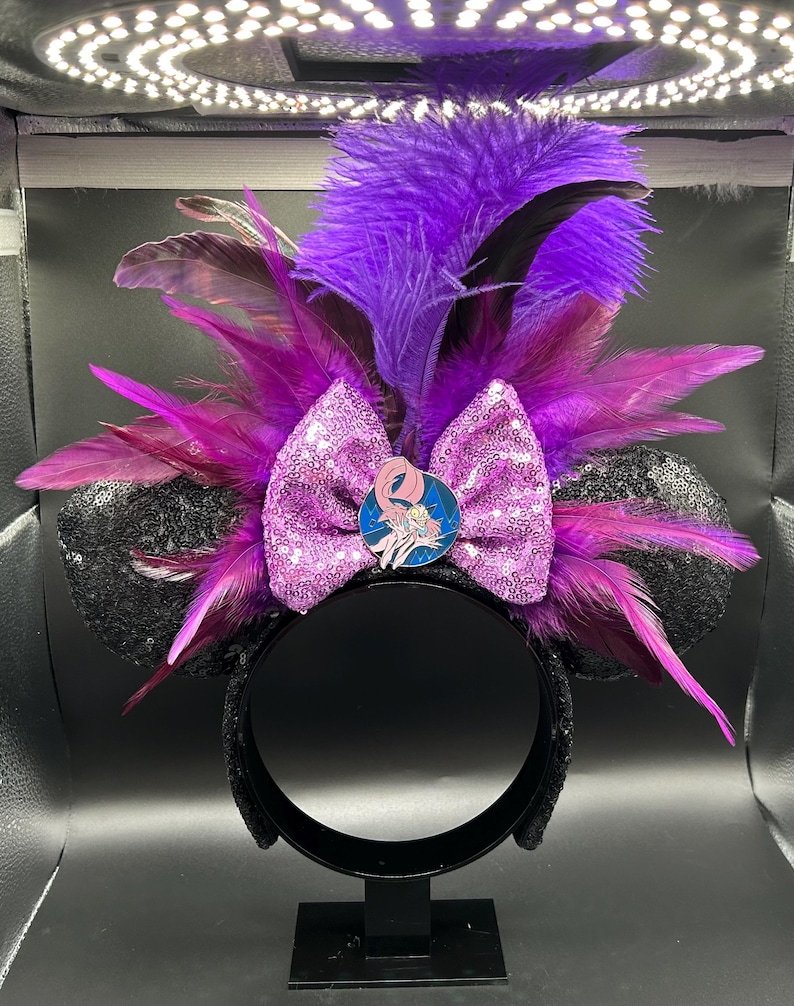 May include: A headband featuring two black sequined ears, a central bright purple sequined bow, and a cluster of vibrant purple and magenta feathers. A circular enamel pin depicting a cartoon character with long hair is centered on the bow.