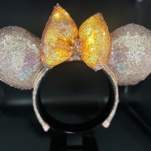 May include: A sequined headband with mouse ears and a bow. The ears and bow are covered in shimmering sequins, with a gold bow and light pink ears. The headband is black.