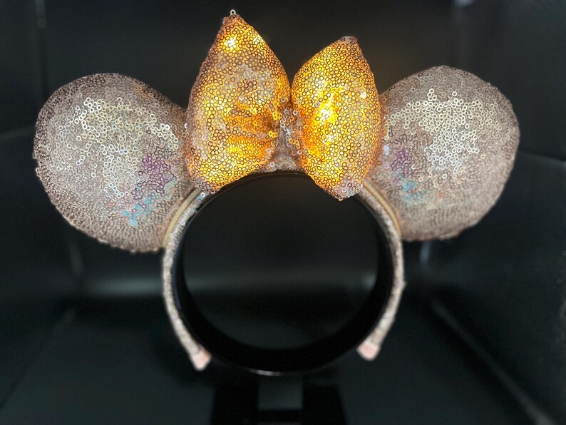 May include: A sequined headband with mouse ears and a bow. The ears and bow are covered in shimmering sequins, with a gold bow and light pink ears. The headband is black.