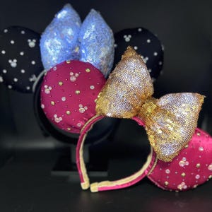 May include: A collection of decorative headbands featuring mouse ears. One headband has black ears with white polka dots, another has blue sequined ears, and a third has magenta ears with a gold sequined bow. All have a black headband.
