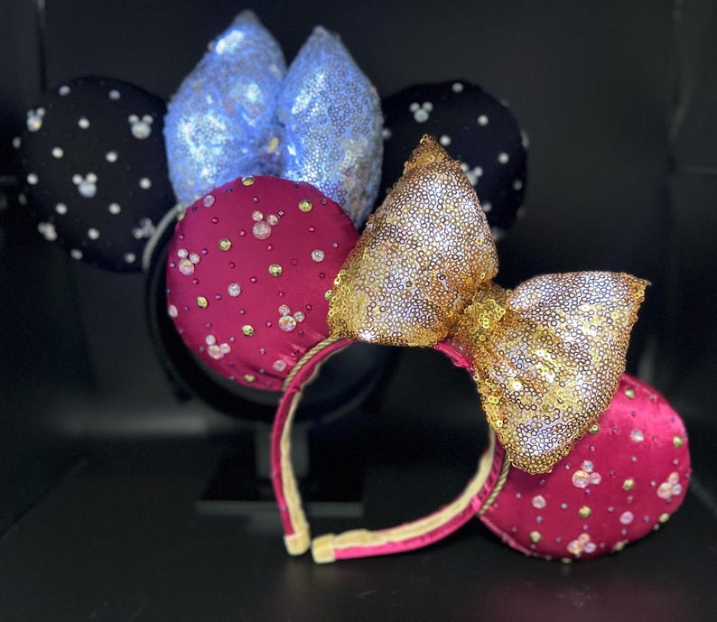 May include: A collection of decorative headbands featuring mouse ears. One headband has black ears with white polka dots, another has blue sequined ears, and a third has magenta ears with a gold sequined bow. All have a black headband.