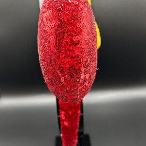 May include: A headband featuring a large, round, red sequined ear. Behind the red ear, a yellow textured element and a white fabric piece are visible. The headband band is also covered in red sequins, providing a consistent look for the accessory.