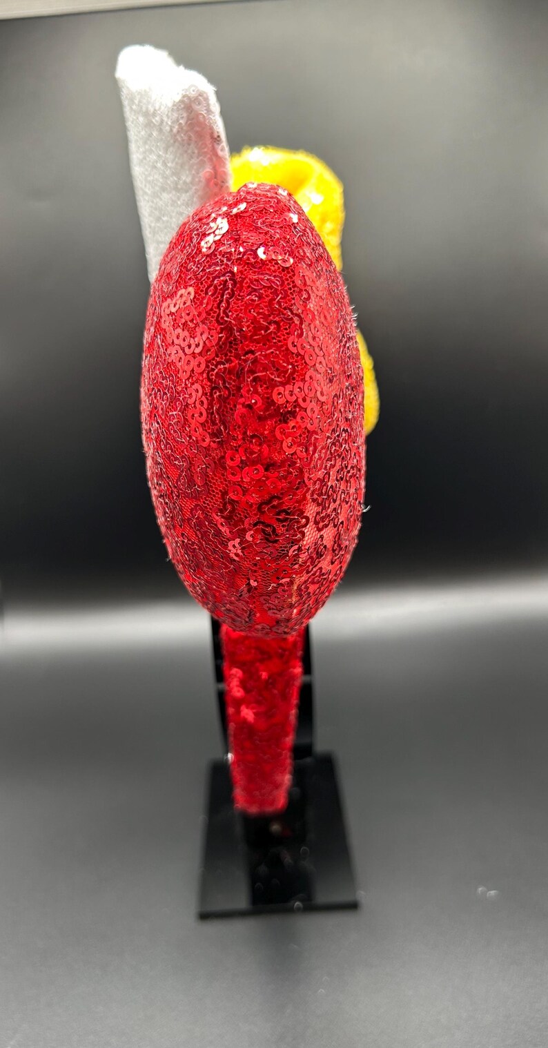 May include: A headband featuring a large, round, red sequined ear. Behind the red ear, a yellow textured element and a white fabric piece are visible. The headband band is also covered in red sequins, providing a consistent look for the accessory.