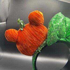 May include: Orange velvet Mickey Mouse ears with green leaf accents and a green sequined bow on a black headband. The ears have a textured, plush appearance, and the bow is covered in shiny sequins.