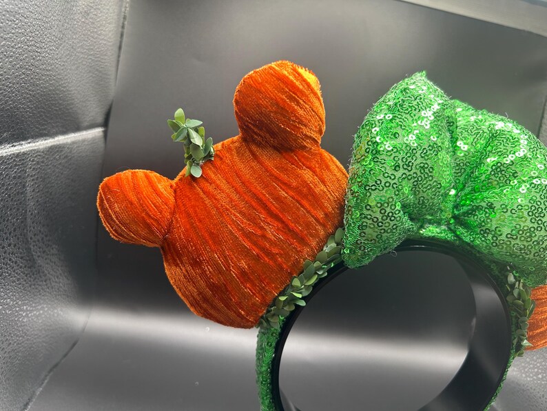 May include: Orange velvet Mickey Mouse ears with green leaf accents and a green sequined bow on a black headband. The ears have a textured, plush appearance, and the bow is covered in shiny sequins.
