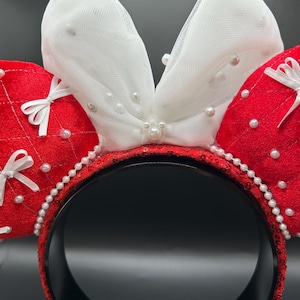 May include: A festive headband featuring red sequined ears with white tulle bows, adorned with faux pearls. The headband itself is black with red sequins and a pearl trim, perfect for themed events.