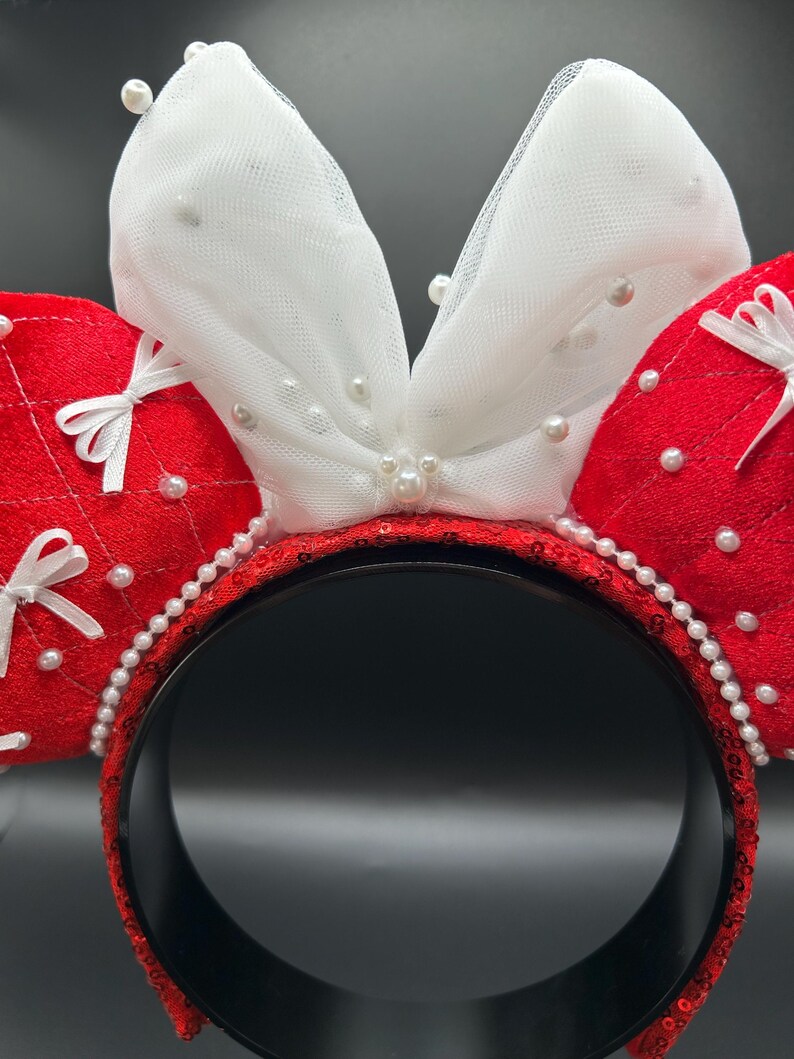 May include: A festive headband featuring red sequined ears with white tulle bows, adorned with faux pearls. The headband itself is black with red sequins and a pearl trim, perfect for themed events.