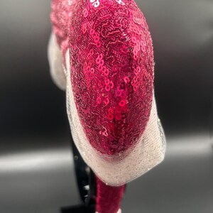 Op de afbeelding: Een roze en witte, met pailletten bezette hoofdband met een groot, hartvormig ontwerp. De hoofdband staat op een zwarte standaard.