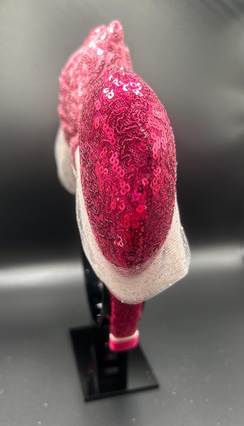 Op de afbeelding: Een roze en witte, met pailletten bezette hoofdband met een groot, hartvormig ontwerp. De hoofdband staat op een zwarte standaard.