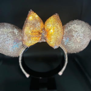 May include: A pair of sequined mouse ears with a gold bow. The ears and headband are covered in shimmering sequins. The bow is a bright gold color, and the ears are a light champagne color.