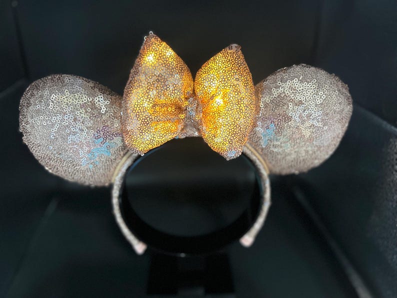 May include: A pair of sequined mouse ears with a gold bow. The ears and headband are covered in shimmering sequins. The bow is a bright gold color, and the ears are a light champagne color.