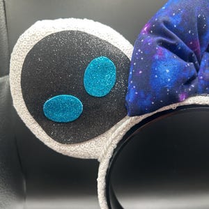 May include: A black headband with white sequined ears and a galaxy-print bow. The ears have a black center with glitter and two turquoise glitter ovals. The bow is a dark blue fabric with a galaxy design.