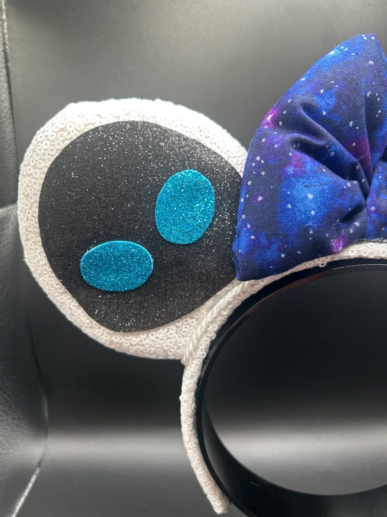 May include: A black headband with white sequined ears and a galaxy-print bow. The ears have a black center with glitter and two turquoise glitter ovals. The bow is a dark blue fabric with a galaxy design.