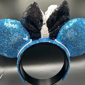 May include: A pair of mouse ear headbands with blue sequined ears and a black sequined bow. The headband is black with a white rope detail. Small white pearls are attached to the ears.