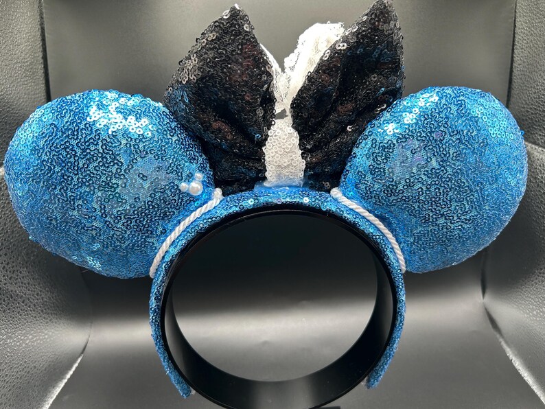 May include: A pair of mouse ear headbands with blue sequined ears and a black sequined bow. The headband is black with a white rope detail. Small white pearls are attached to the ears.