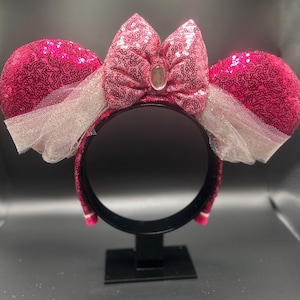 Op de afbeelding: Roze glitzerende Minnie Mouse oren met een grote roze strik en witte tule. De oren staan op een zwarte standaard.