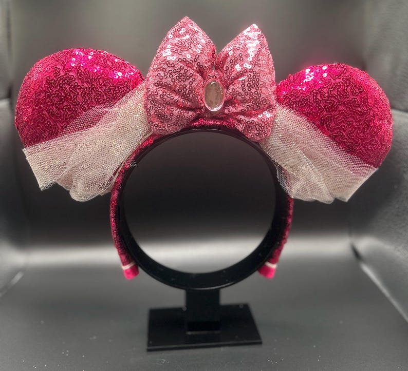 Op de afbeelding: Roze glitzerende Minnie Mouse oren met een grote roze strik en witte tule. De oren staan op een zwarte standaard.