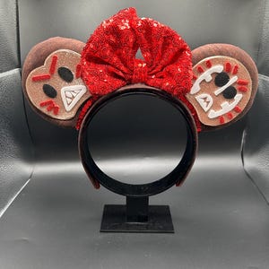 May include: A headband featuring two round brown ears with gingerbread-inspired face appliqu&eacute;s in white, black, and red. A large, bright red sequined bow sits between the ears. The headband frame is dark brown and has a smooth, solid finish.