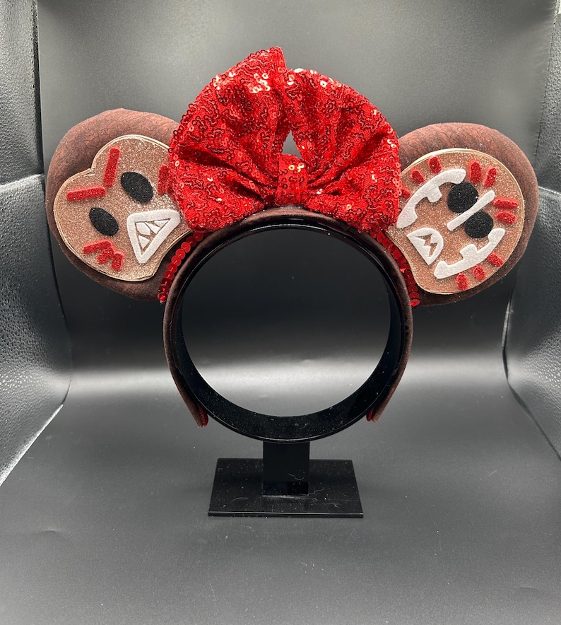 May include: A headband featuring two round brown ears with gingerbread-inspired face appliqu&eacute;s in white, black, and red. A large, bright red sequined bow sits between the ears. The headband frame is dark brown and has a smooth, solid finish.