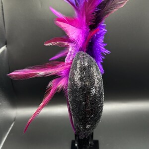May include: A headband featuring a black sequined ear-shaped component adorned with a large, upright arrangement of vibrant purple and magenta feathers. The feathers extend vertically and horizontally from the side of the sequined base.