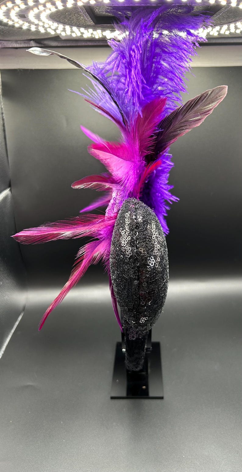May include: A headband featuring a black sequined ear-shaped component adorned with a large, upright arrangement of vibrant purple and magenta feathers. The feathers extend vertically and horizontally from the side of the sequined base.