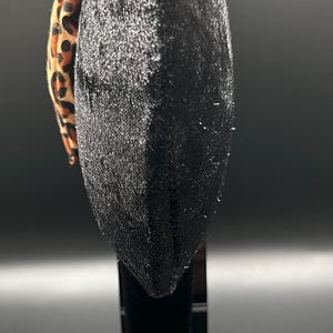 以下が含まれることがあります： 黒いベルベットのヘアバンドとレオパード柄のリボン。ヘアバンドは、暗い背景の黒いスタンドに展示されています。レオパード柄は茶色と黒の斑点があります。