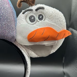 May include: A white sequined headband featuring a plush Olaf character from the movie Frozen. The Olaf character has a carrot nose, black eyes, and brown twig-like hair. The headband is black.
