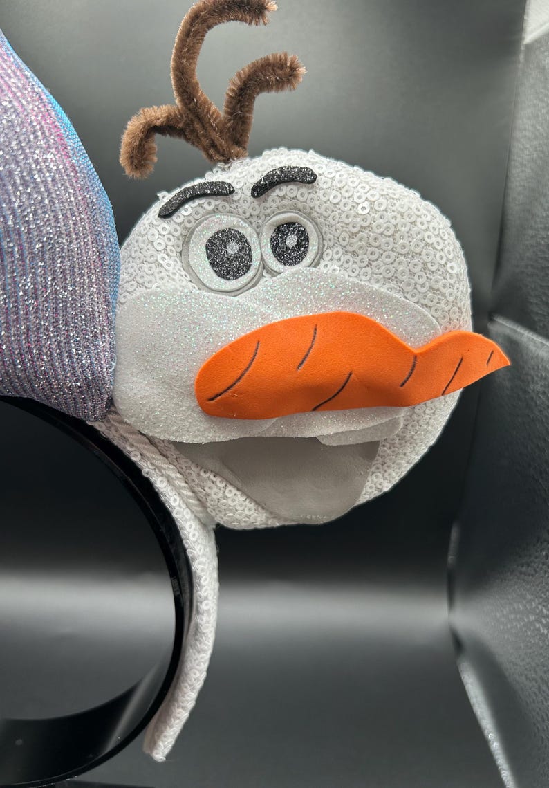 May include: A white sequined headband featuring a plush Olaf character from the movie Frozen. The Olaf character has a carrot nose, black eyes, and brown twig-like hair. The headband is black.