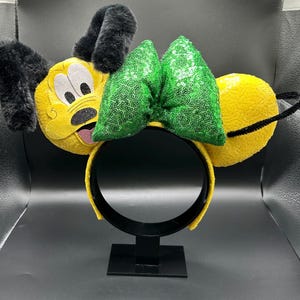 May include: A yellow headband with plush dog ears and a green sequined bow. The dog's face is embroidered on the front of the headband, with black ears and a black nose. The headband is on a black stand.
